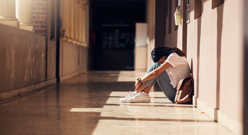 En flicka sitter ensam i en skolkorridor. Hon har knäna uppdragna och lutar huvudet mot knäna. 