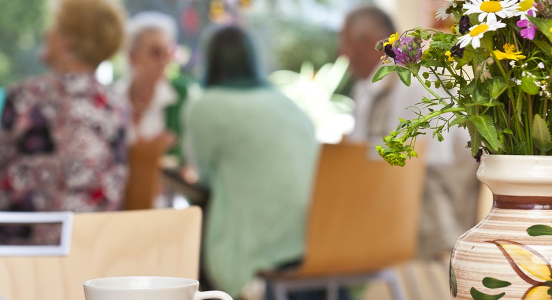 Äldre sitter runt ett bord och fikar. I förgrunden en kaffekopp och blommor