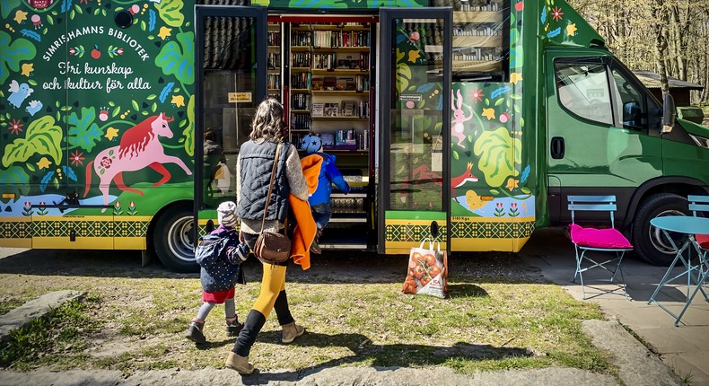 Bokbussen står parkerad i nationalparken Stenshuvud mot en fond av träd. En förälder med barn är på väg in i bokbussen.