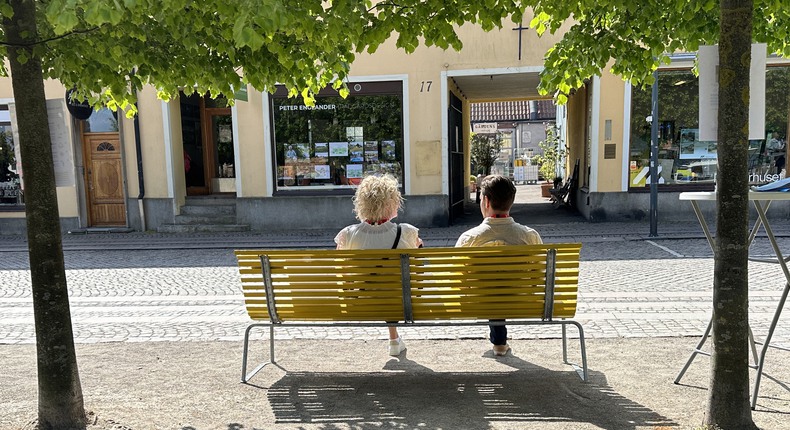 Två personer sitter på en gul bänk under grönskande träd.