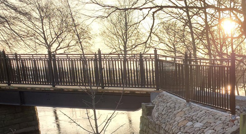 En bro med svarta metallräcken som går över Tommarpsån. I bakgrunden lyser solen genom träd med bara grenar.