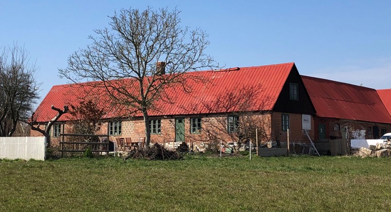 Flera husbyggnationer av sten och tegel med rött tak. Boningshuset har gröna fönster och dörrar. Framför huset står träd och en flaggstång.